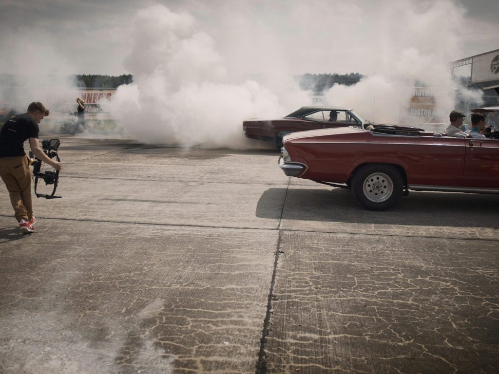 A dynamic scene of classic cars and filmmakers capturing a smoky drag race event.