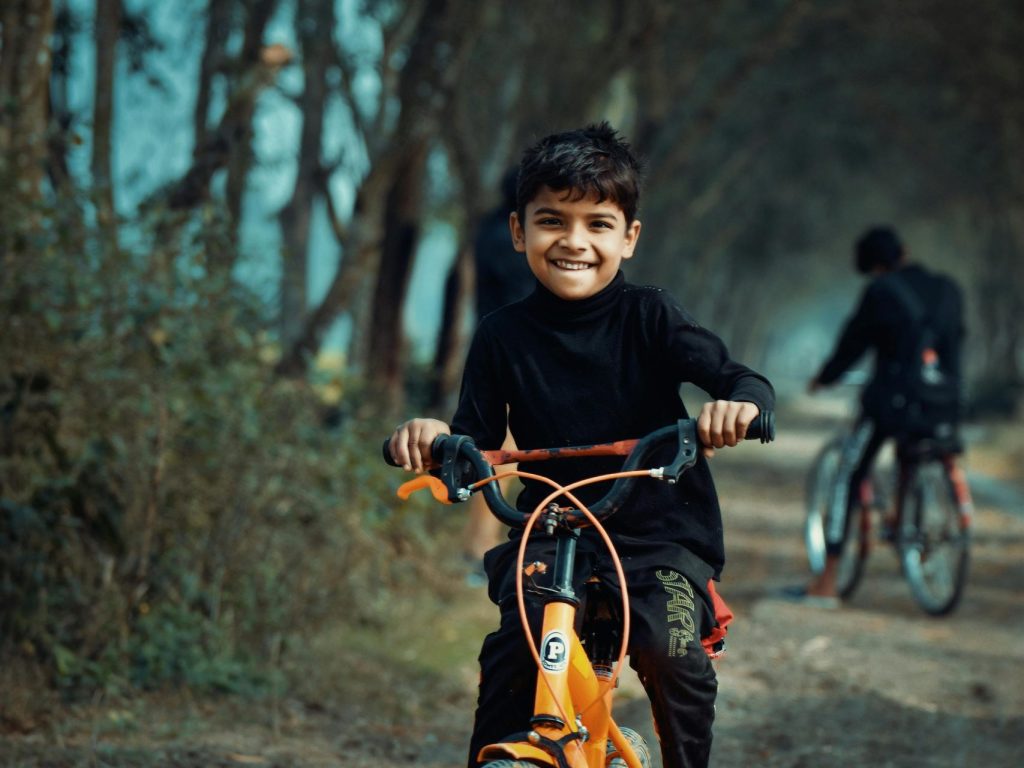 Happy child rides an orange bicycle on a rural dirt path surrounded by trees in Bangladesh.